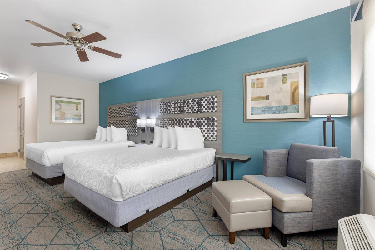 A hotel room with two beds, a ceiling fan, a patterned wall, a small table, a sofa, and a lamp.