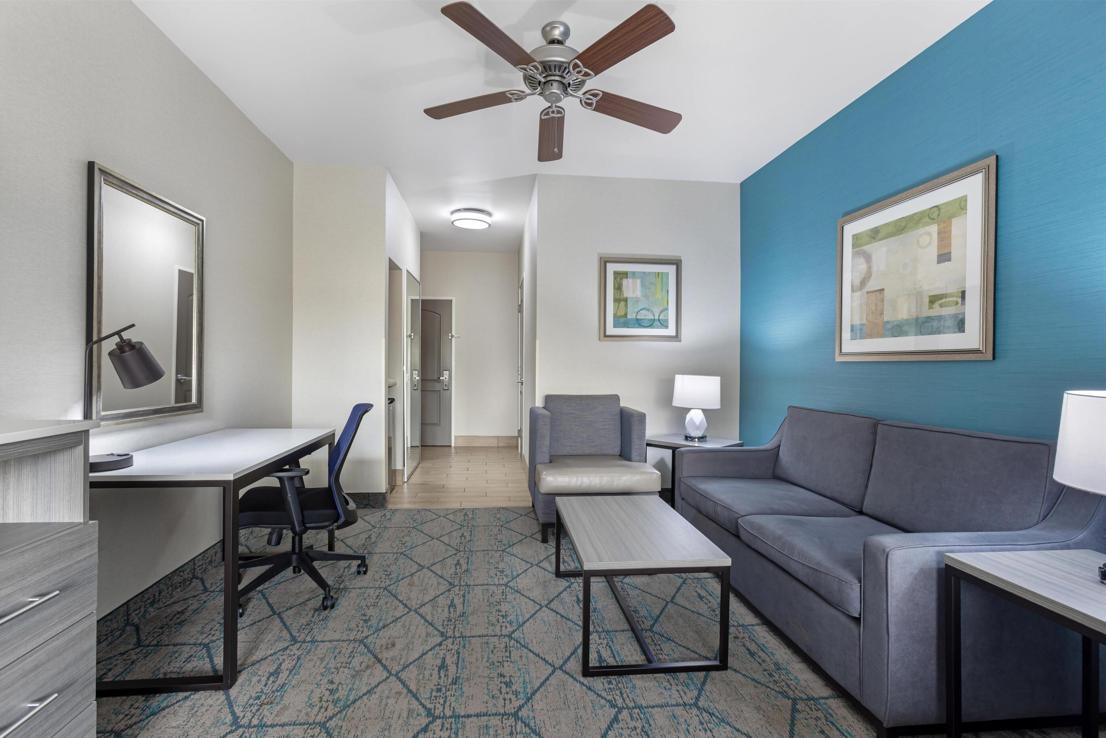 A hotel room with a desk, a sofa, a chair, a ceiling fan, a framed picture, and a patterned carpet.
