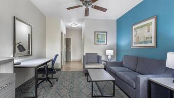 A hotel room with a desk, a sofa, a chair, a ceiling fan, a framed picture, and a patterned carpet.