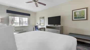 A hotel room with a large bed, a desk with a chair, a television, a ceiling fan, a window, and a painting on the wall.