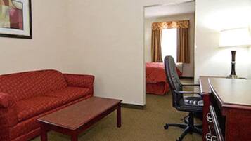 Habitación de hotel con sofá rojo, una mesita de centro de madera, una silla de oficina negra, un escritorio con lámpara y una cama con cubierta roja.