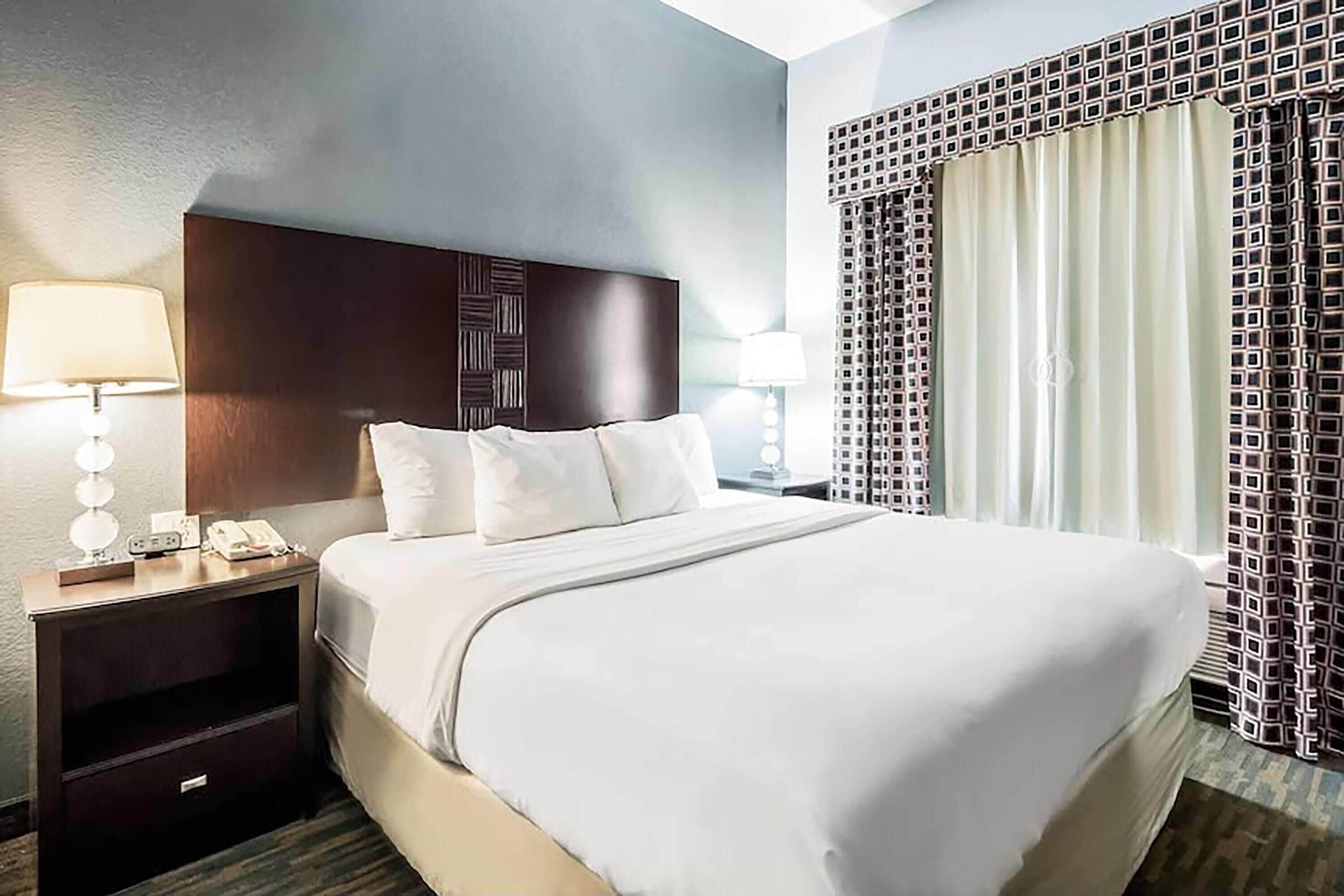 A hotel room with a large bed, a nightstand with a lamp, a telephone, and a patterned curtain.
