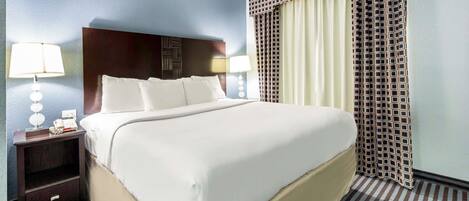 A hotel room with a large bed, a nightstand with a lamp, and a window with patterned curtains.