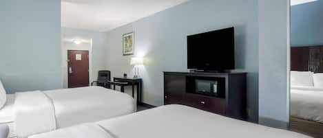 A hotel room with two beds, a TV on a stand, a desk with a lamp, and a door in the background.