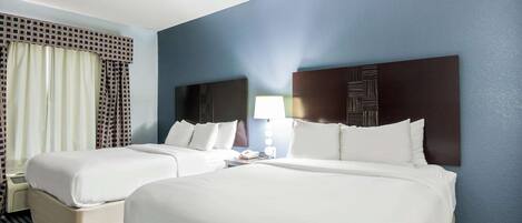 A hotel room with two beds, a large window with curtains, a bedside table with a lamp, and a wall-mounted artwork.