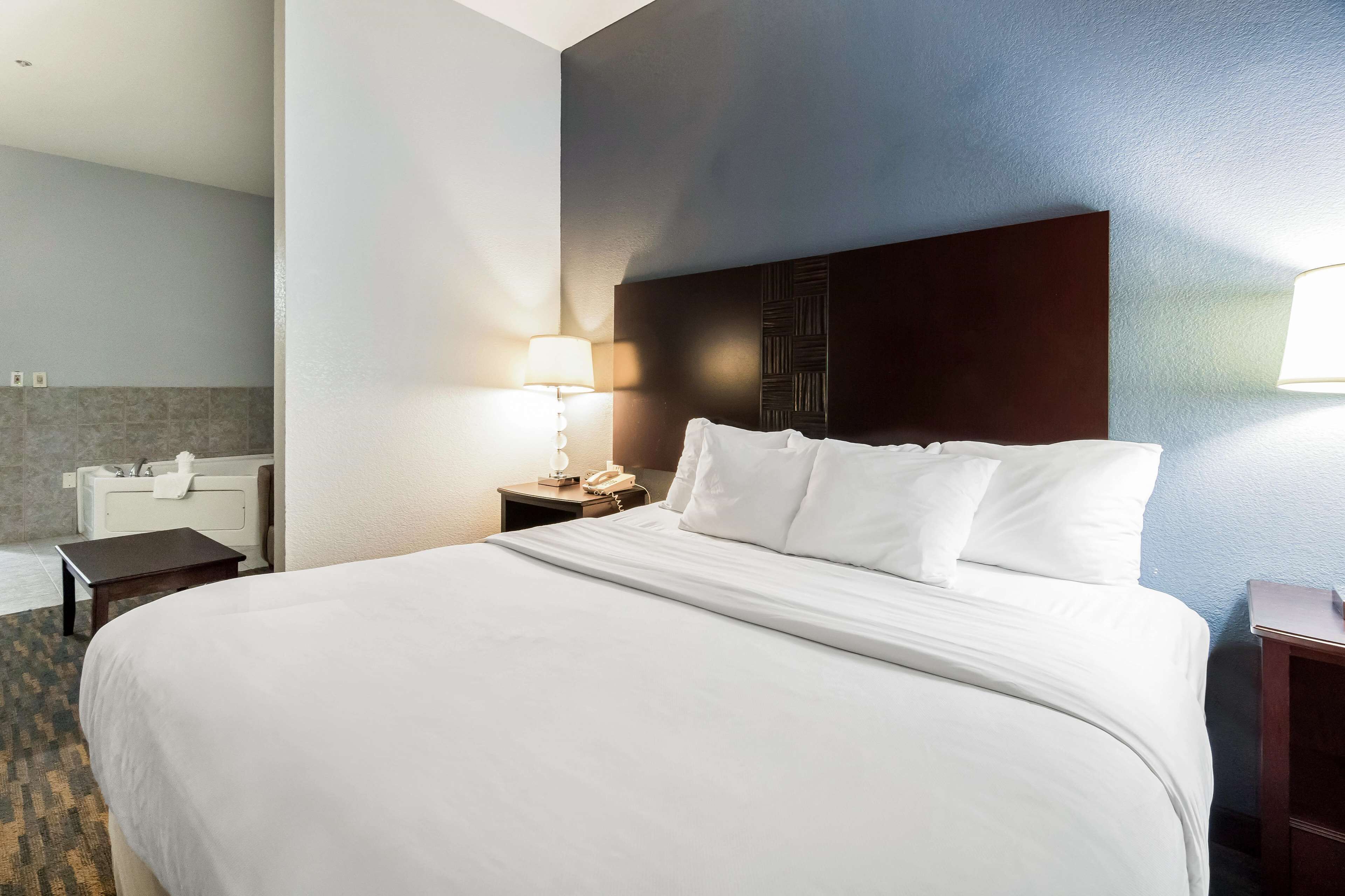 A hotel room with a large bed, a bedside table with a lamp, a small table, and a bathroom with a bathtub and sink.