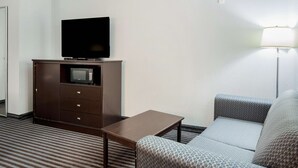 A hotel room with a flat-screen TV on a wooden cabinet, a microwave, a small table, a grey patterned sofa, and a floor lamp. - Baymont by Wyndham Mineral Wells (Mineral Wells)