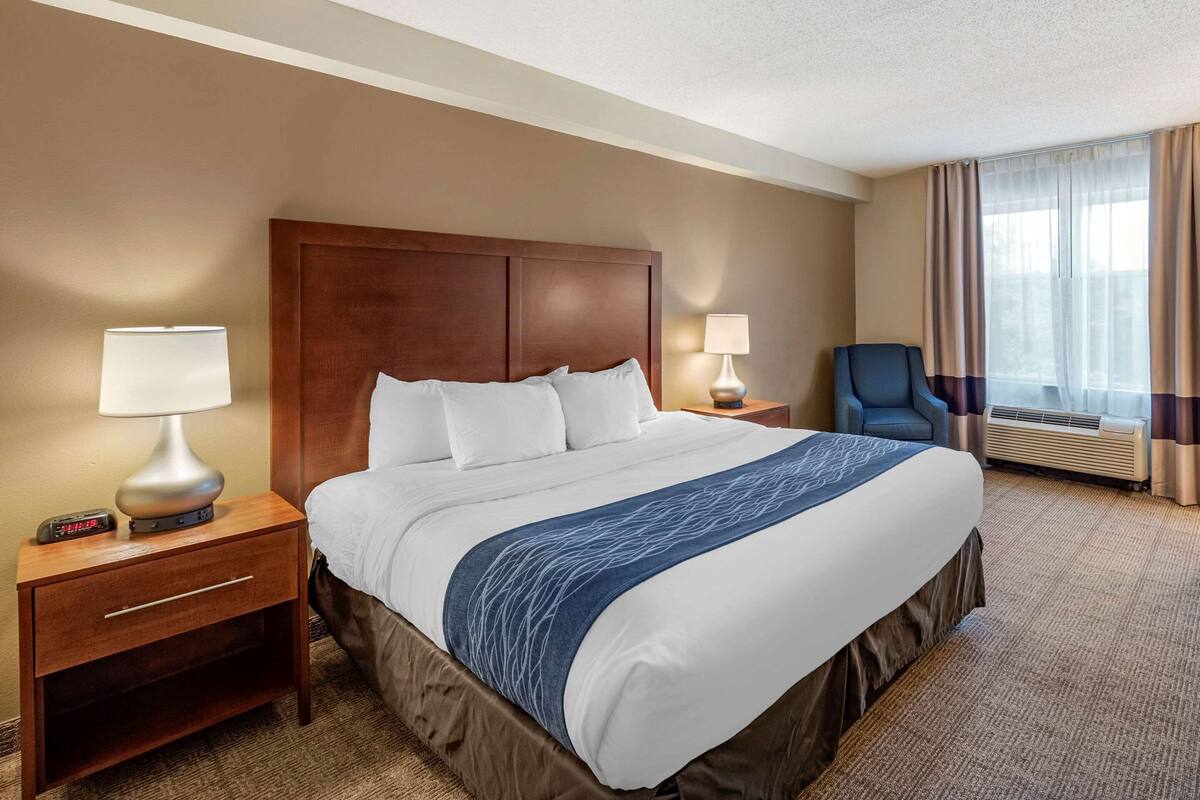 Una habitación de hotel con una cama grande, dos mesitas de noche con lámparas, una silla y un ventanal con cortinas.