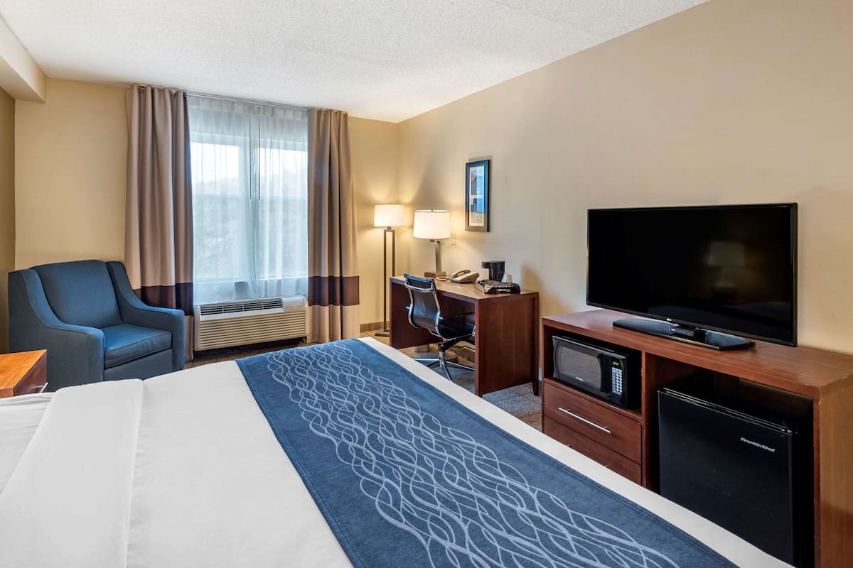 Habitación de hotel con cama, escritorio, televisor, mini refrigerador, silla y ventana con cortinas.