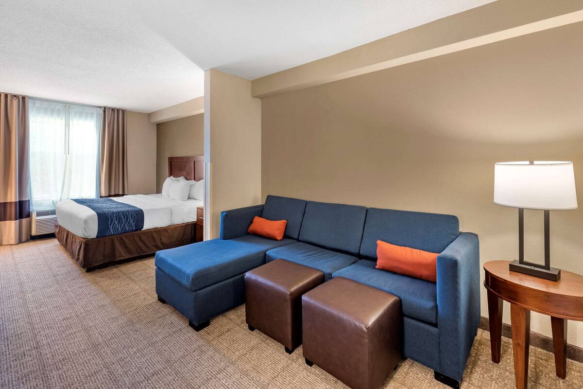 Habitación de hotel con cama, sofá, dos otomanas, lámpara y ventana con cortinas.