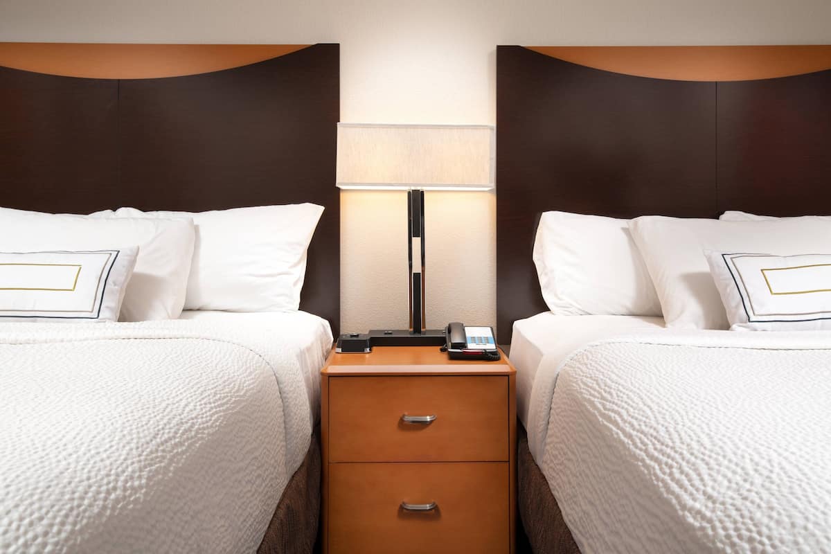 A hotel room with two beds, a nightstand with a lamp, and a phone.