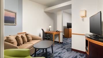 A hotel room with a sofa, a small table, a green chair, a desk with a lamp, a television, and a carpeted floor.