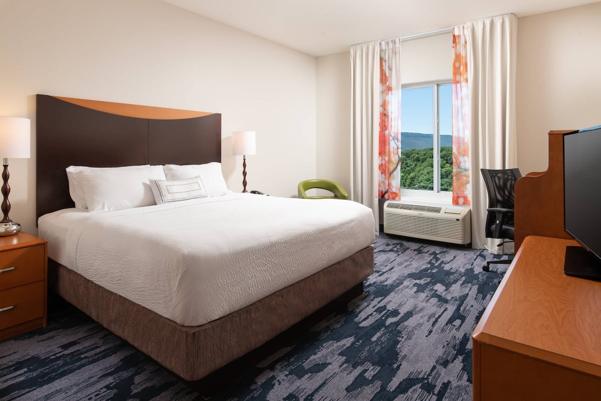 A hotel room with a large bed, a desk with a television, a chair, a desk lamp, and a window with curtains.
