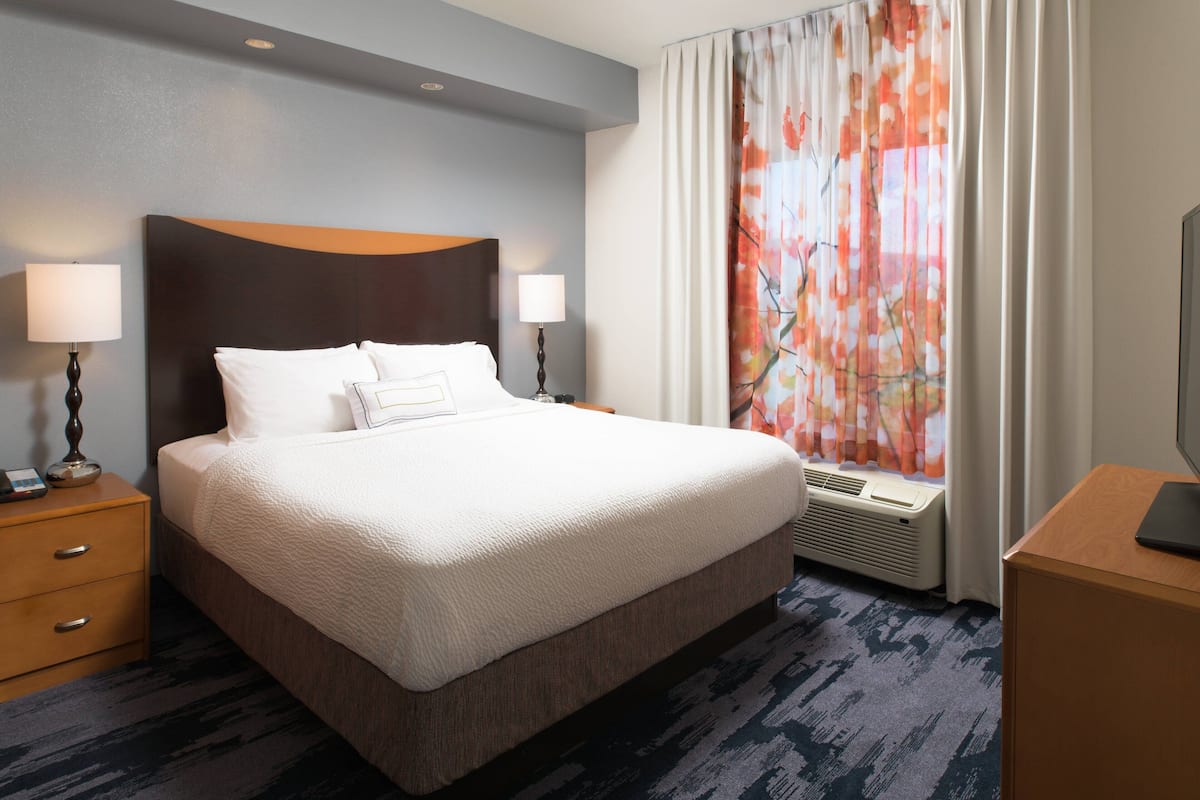 A hotel room with a large bed, two bedside tables with lamps, a window with curtains, and a desk with a television.
