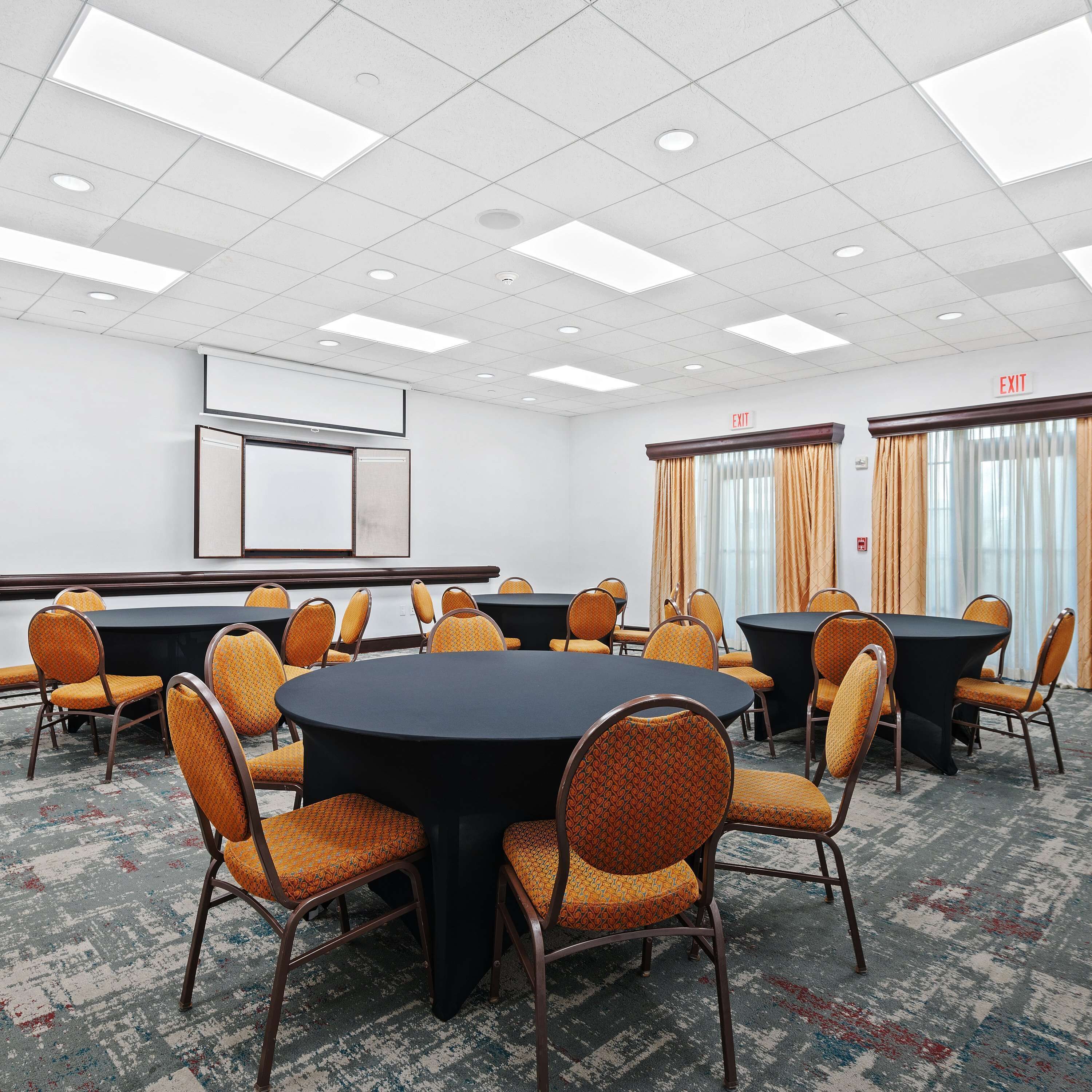A conference room with multiple tables and chairs, a projector screen, and an exit sign.