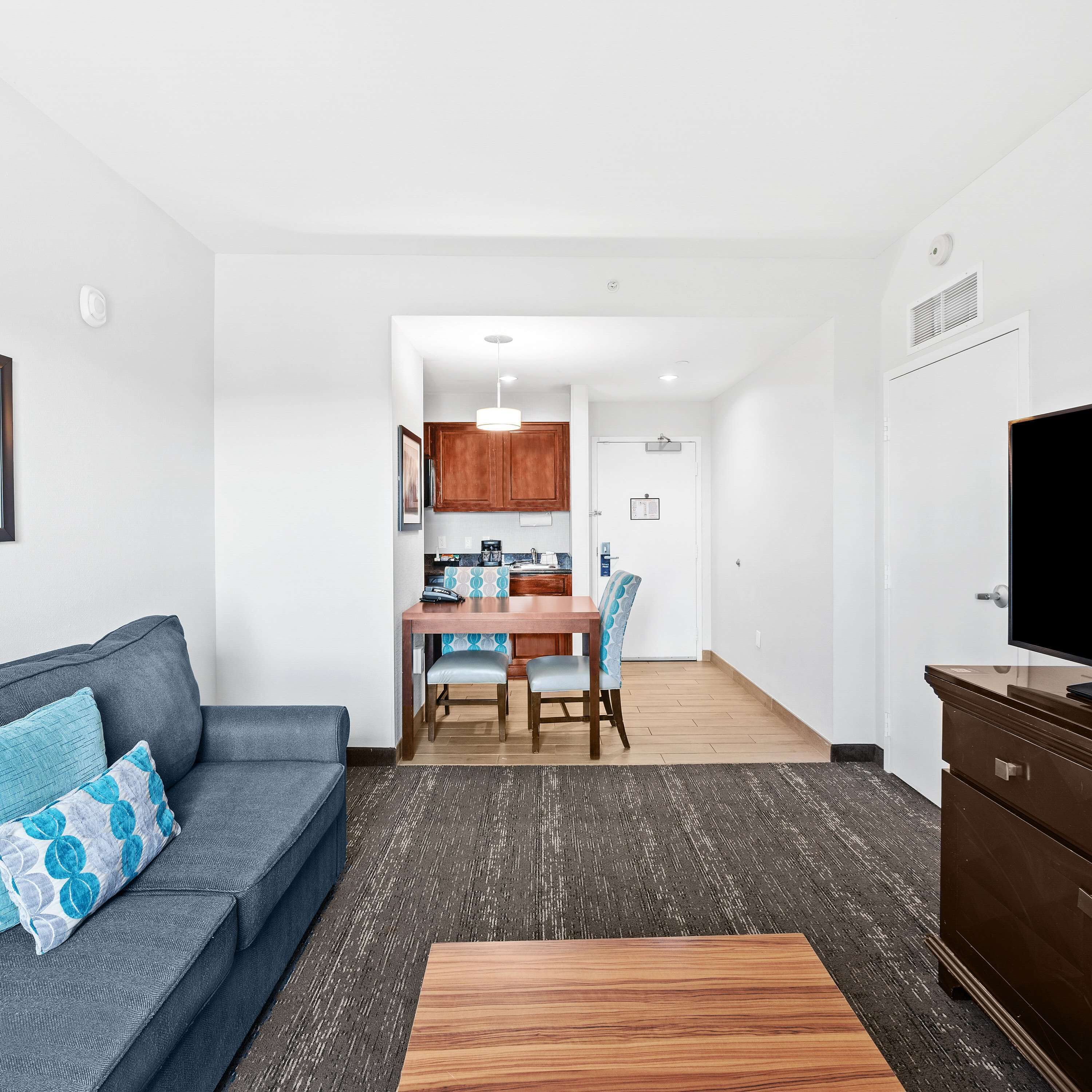 A modern living room with a sofa, a coffee table, and a TV. There is a dining area with a table and chairs.