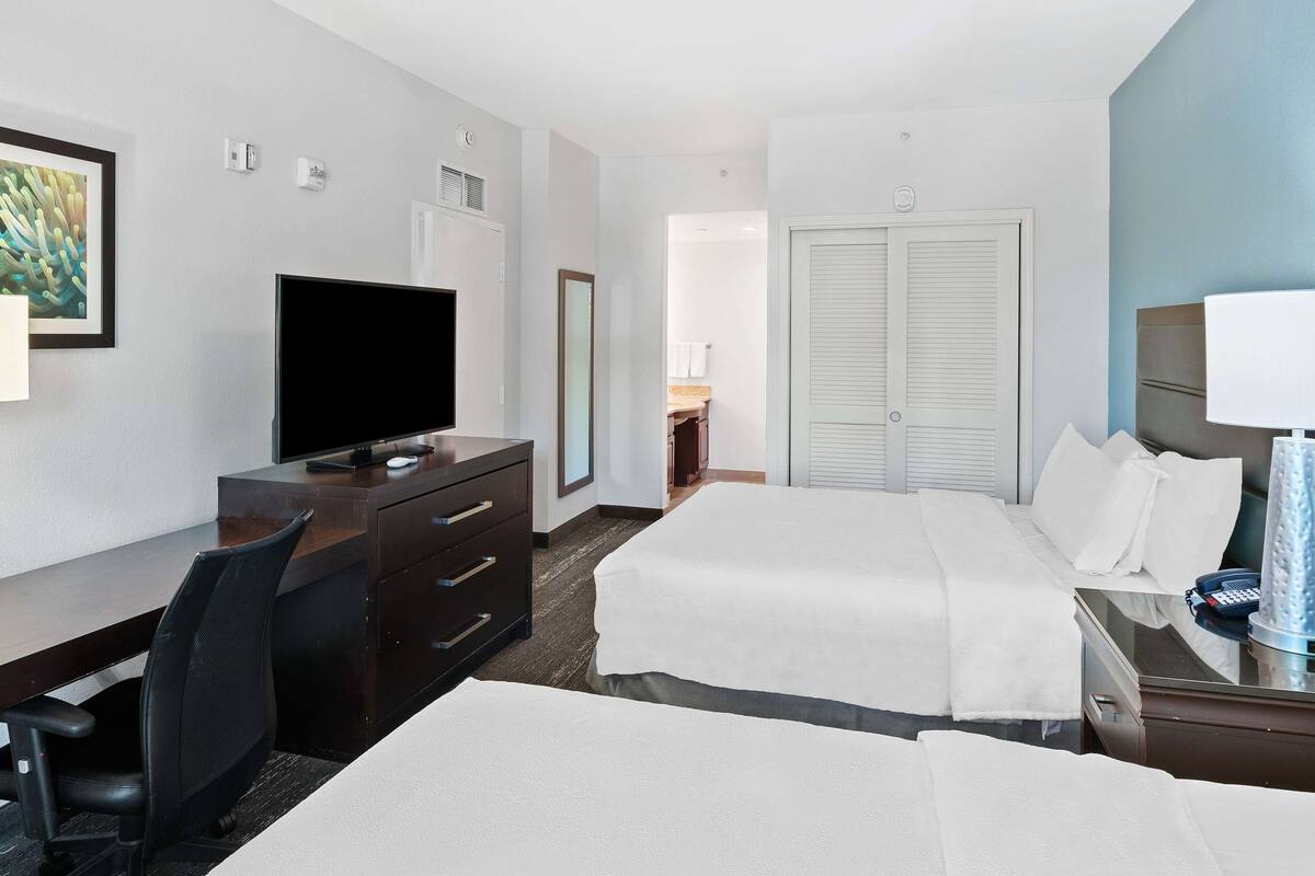Una camera d'albergo con una scrivania, due letti, una televisione, uno specchio e una porta.