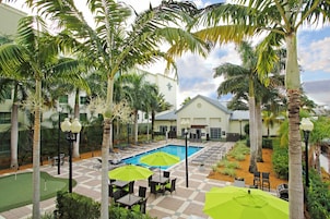 A pool area with lounge chairs and umbrellas, surrounded by palm trees and a building with a white facade.