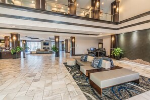 A spacious lobby with a high ceiling, a seating area with sofas and a coffee table, a reception desk, and a large TV screen.