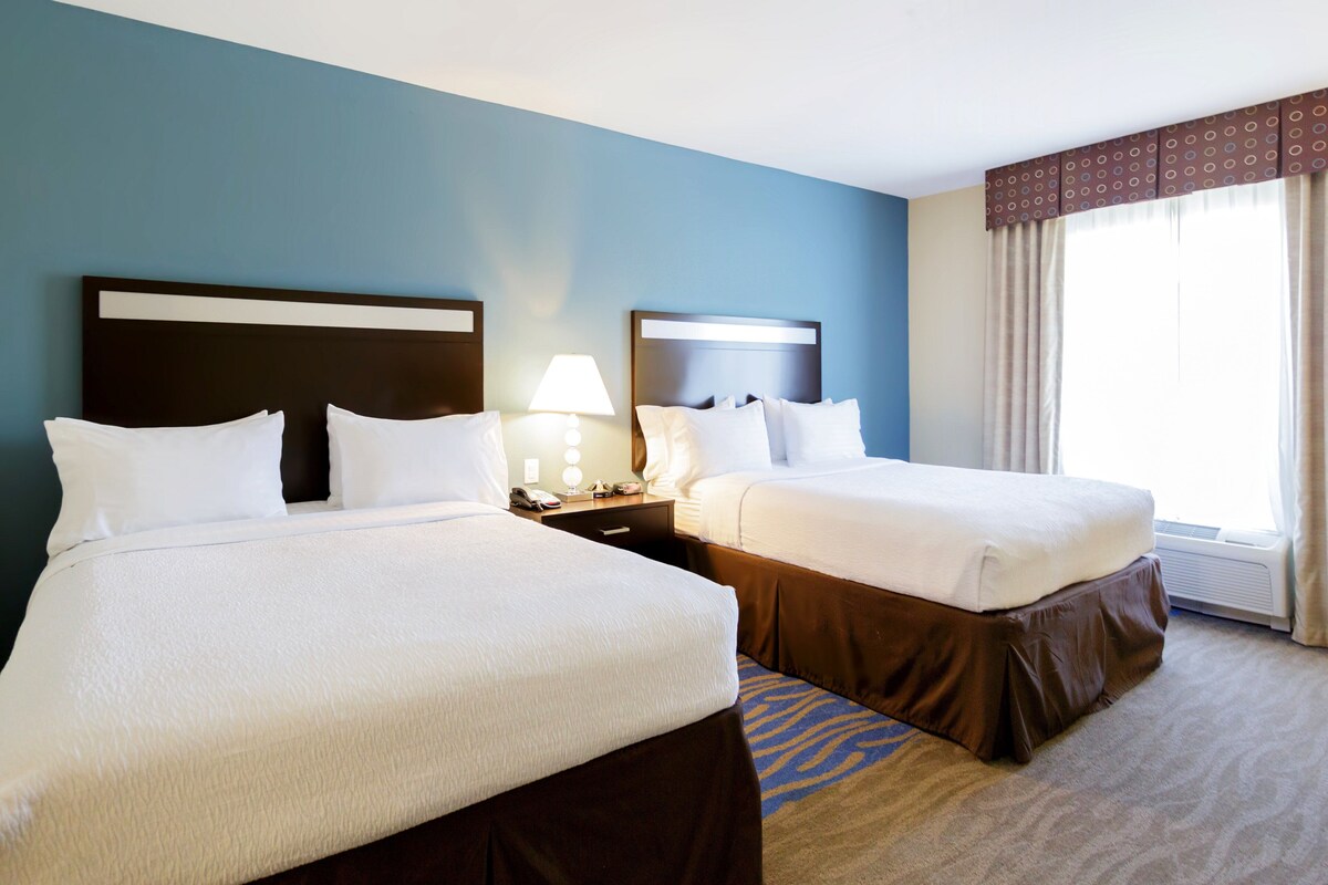 A hotel room with two beds, a nightstand with a lamp, a window with curtains, and a carpeted floor.