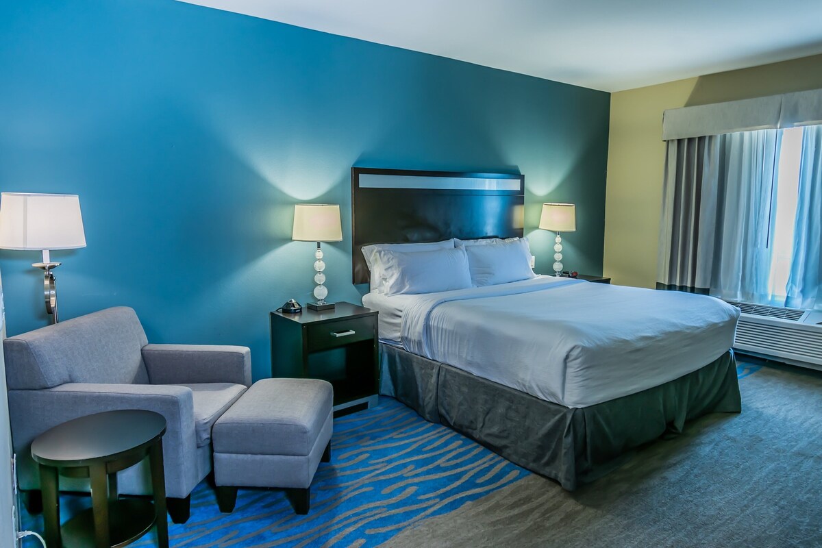 A hotel room with a bed, bedside lamps, a chair, a small table, and a window with curtains.
