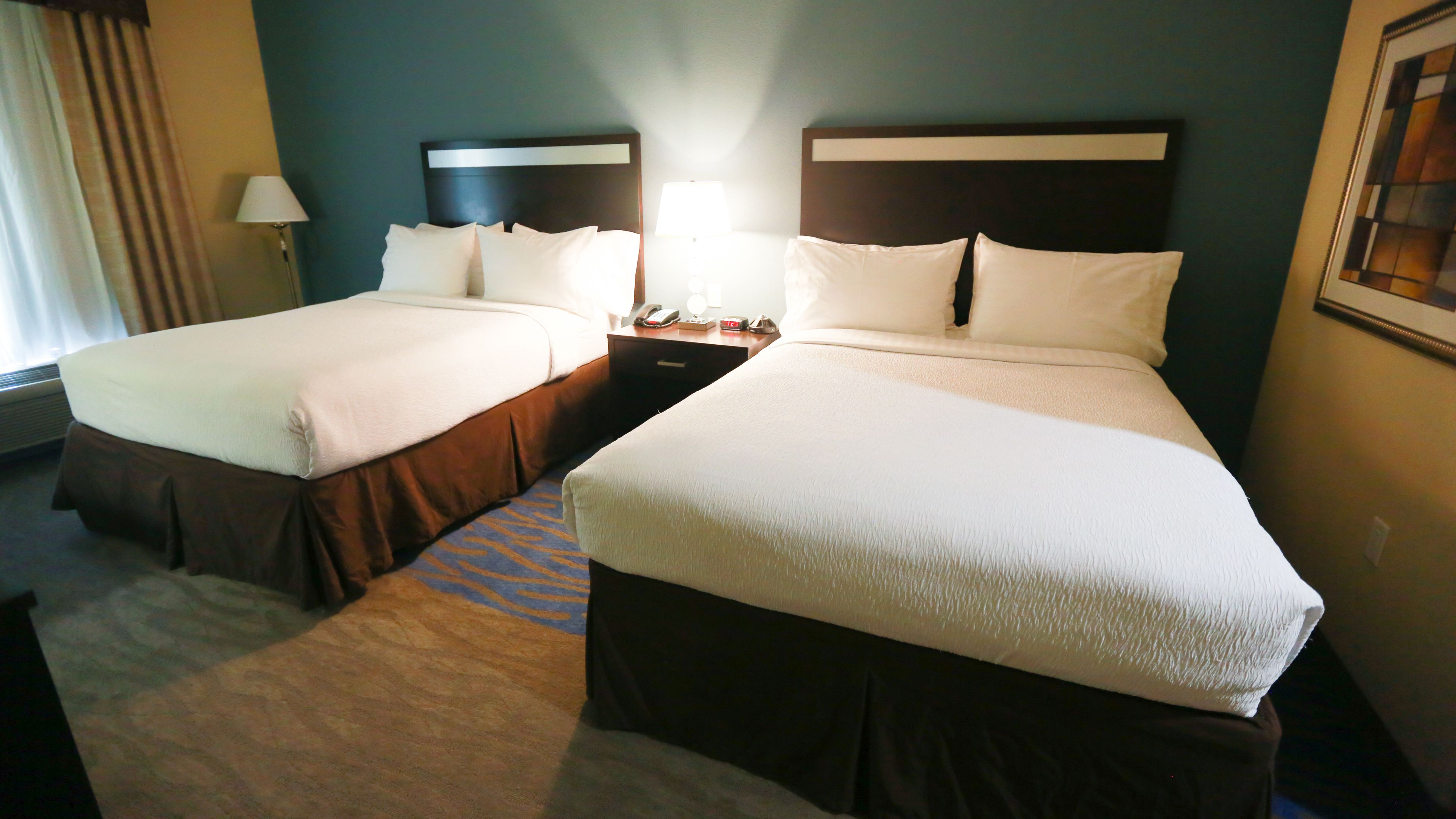 A hotel room with two beds, a nightstand with a lamp, and a window with curtains.
