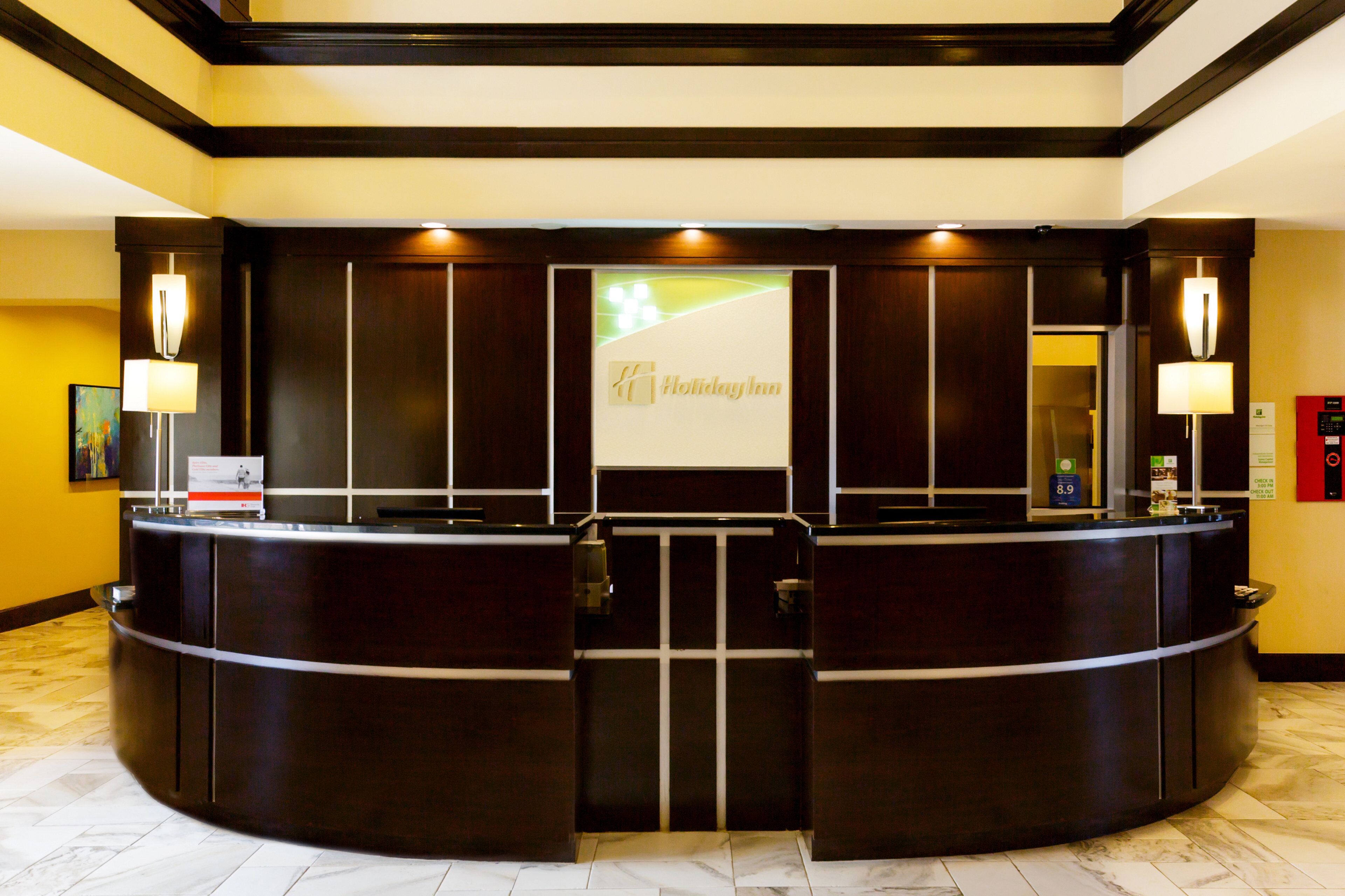 Mostrador de recepción de hotel con pantalla digital mostrando el nombre del hotel, un mostrador de registro y una zona de recepción con suelo de mármol.