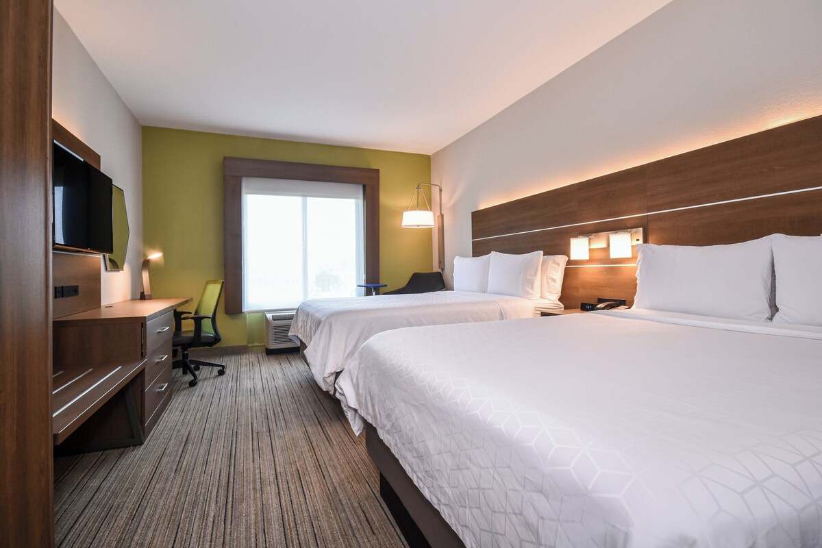 Habitación de hotel con dos camas, un escritorio con silla, un televisor y una ventana con persianas.