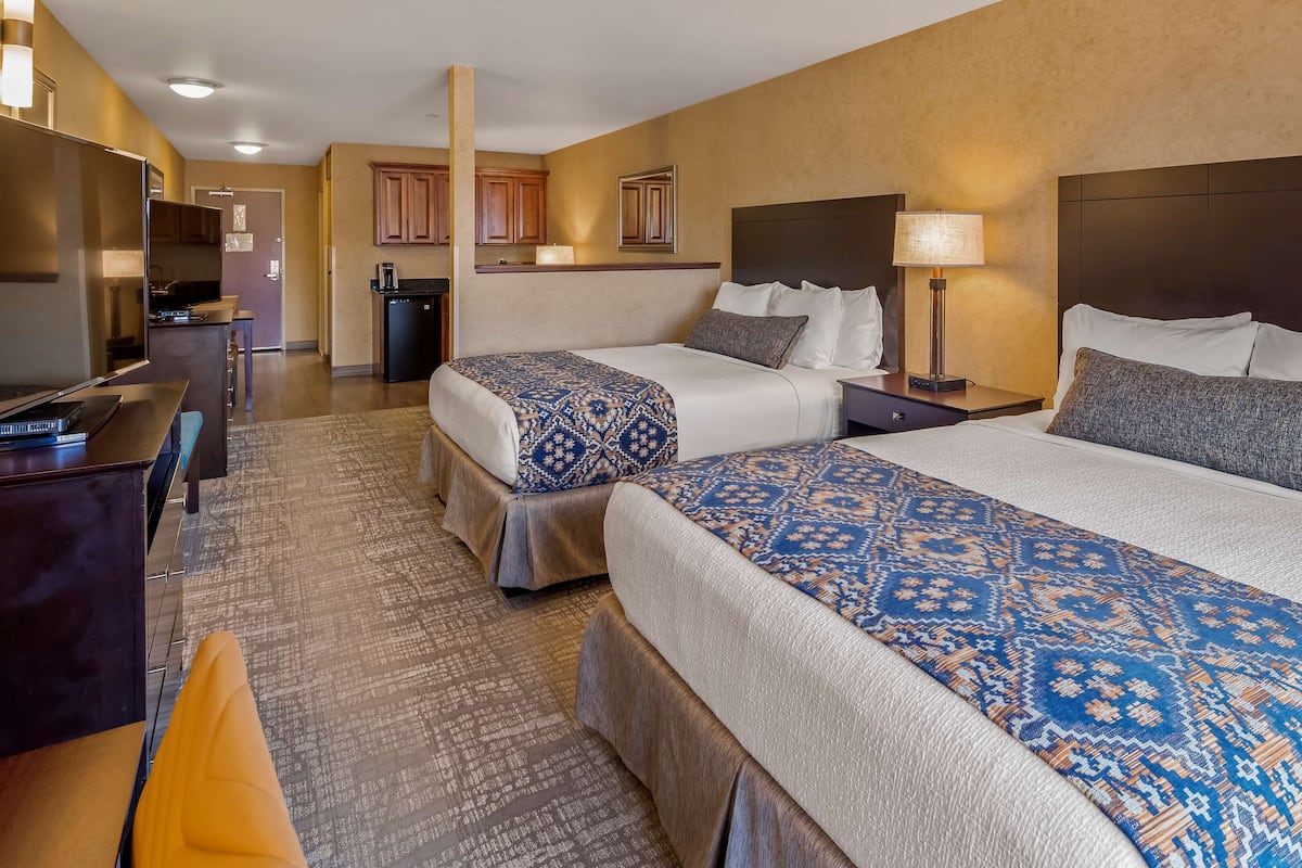 A hotel room with two beds, a desk, a chair, a small kitchenette, and a bathroom.