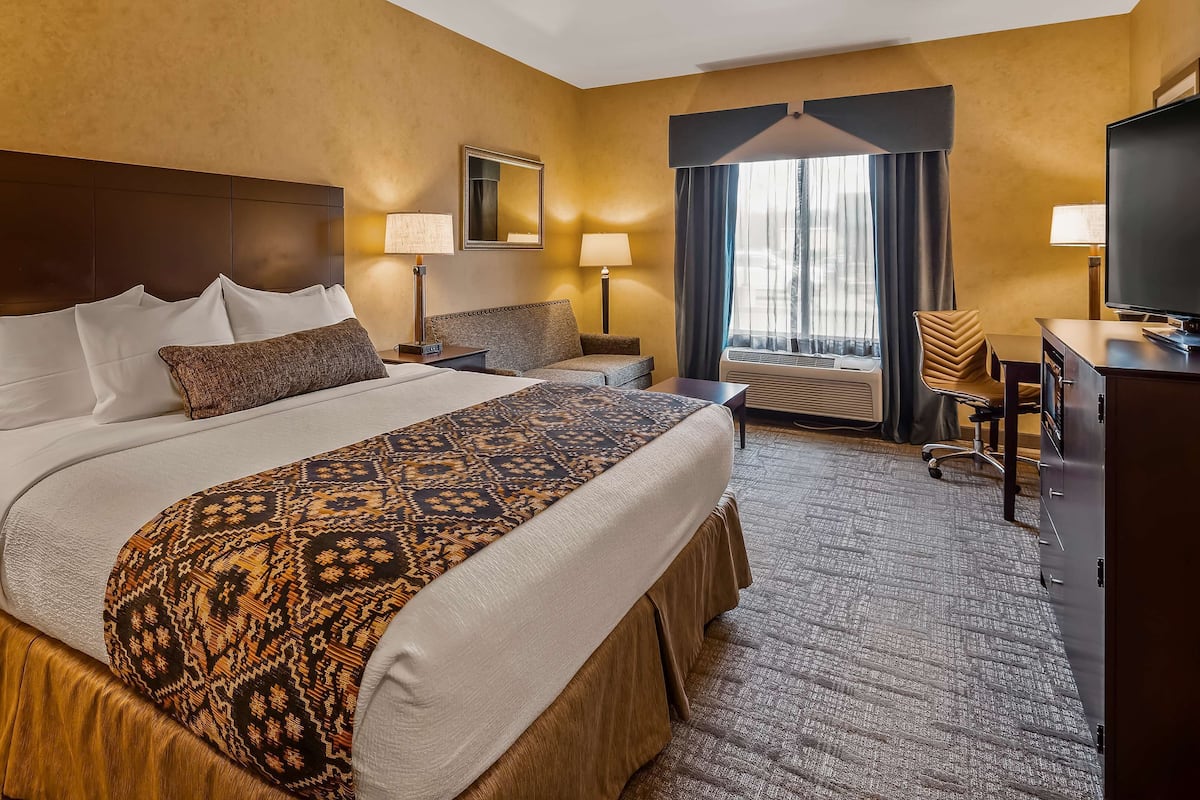 A hotel room with a large bed, two bedside tables with lamps, a desk with a chair, a large mirror, and a window with curtains.