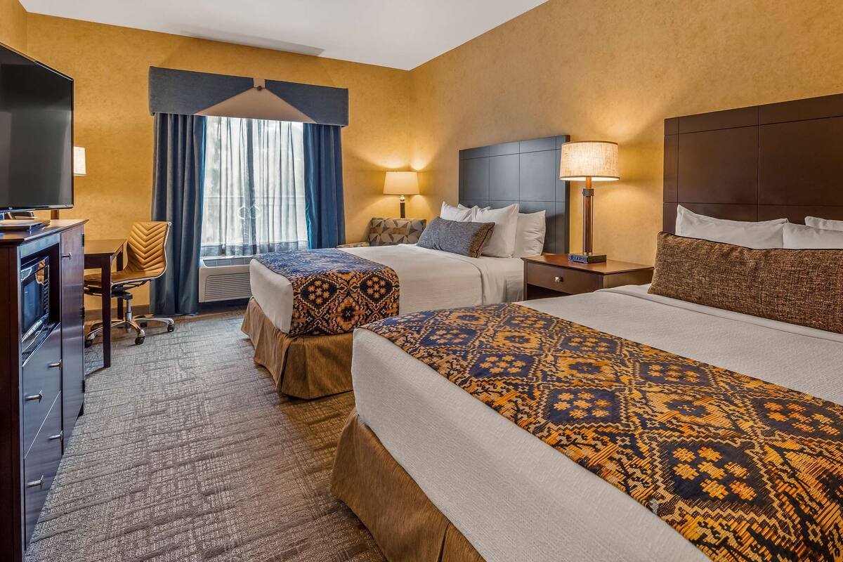 A hotel room with two beds, a desk with a flat-screen TV, a chair, and a window with curtains.