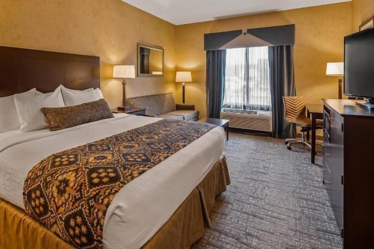 A hotel room with a large bed, two bedside tables, a desk, a chair, a window with curtains, and a mirror on the wall.