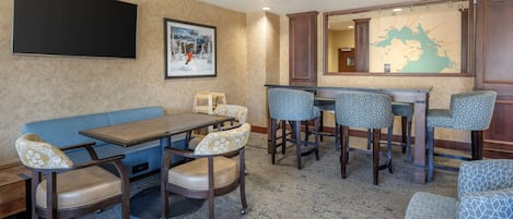 En hotellobby med en stor platt-TV, en karta på väggen, en blå soffa och en bardisk med mönstrade barstolar.