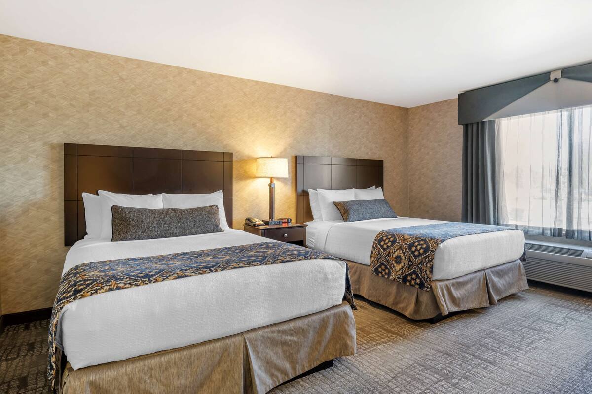 A hotel room with two beds, a nightstand with a lamp, and a large window with curtains.