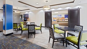 Un hall de hotel con mostrador de recepción, zona de estar con mesas y sillas, y vista al área de la cocina.
