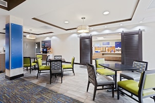 Un hall de hotel con mostrador de recepción, zona de estar con mesas y sillas, y vista al área de la cocina.