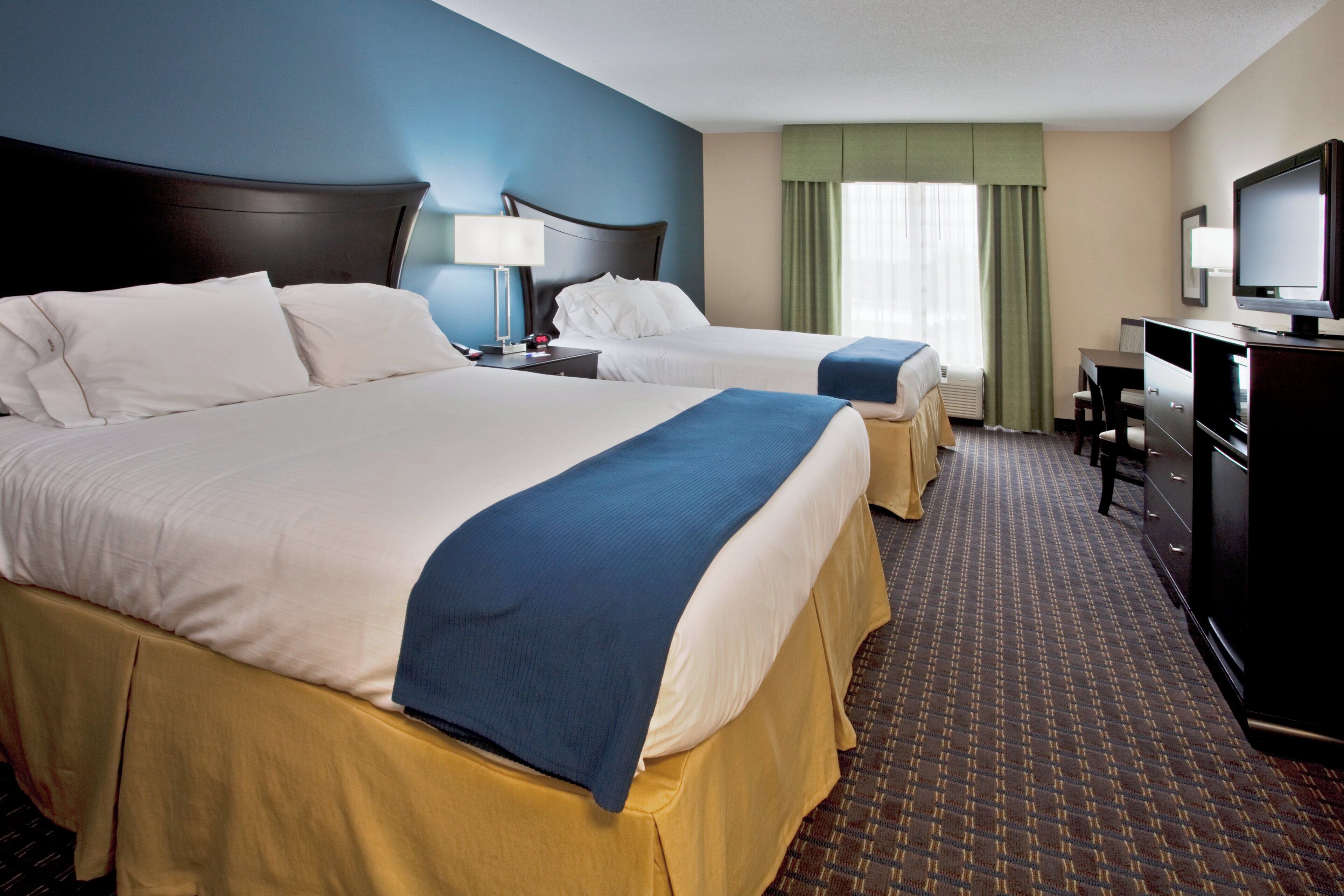 A hotel room with two beds, a desk, a television, and a window with curtains.