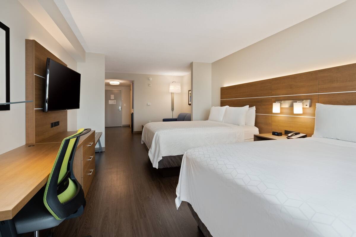 A hotel room with two beds, a desk with a chair, a flat-screen TV mounted on the wall, and a bedside table with lamps.