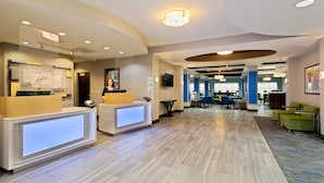A modern hotel lobby with a reception desk, seating area, and a staircase leading to an elevator.