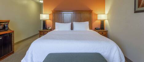A hotel room with a large bed, two bedside tables with lamps, a wooden headboard, a small desk with a telephone, and a bench at the foot of the bed.