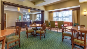 Un vestíbulo de hotel con comedor que presenta mesas y sillas de madera, una cocina al fondo y amplias ventanas con cortinas.