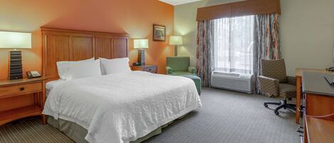 A hotel room with a large bed, two bedside tables, a desk with a chair, a window with curtains, and a radiator.