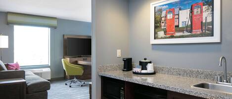 A modern hotel room with a kitchenette, a gray sofa, a green chair, and a painting of red gas pumps on the wall.