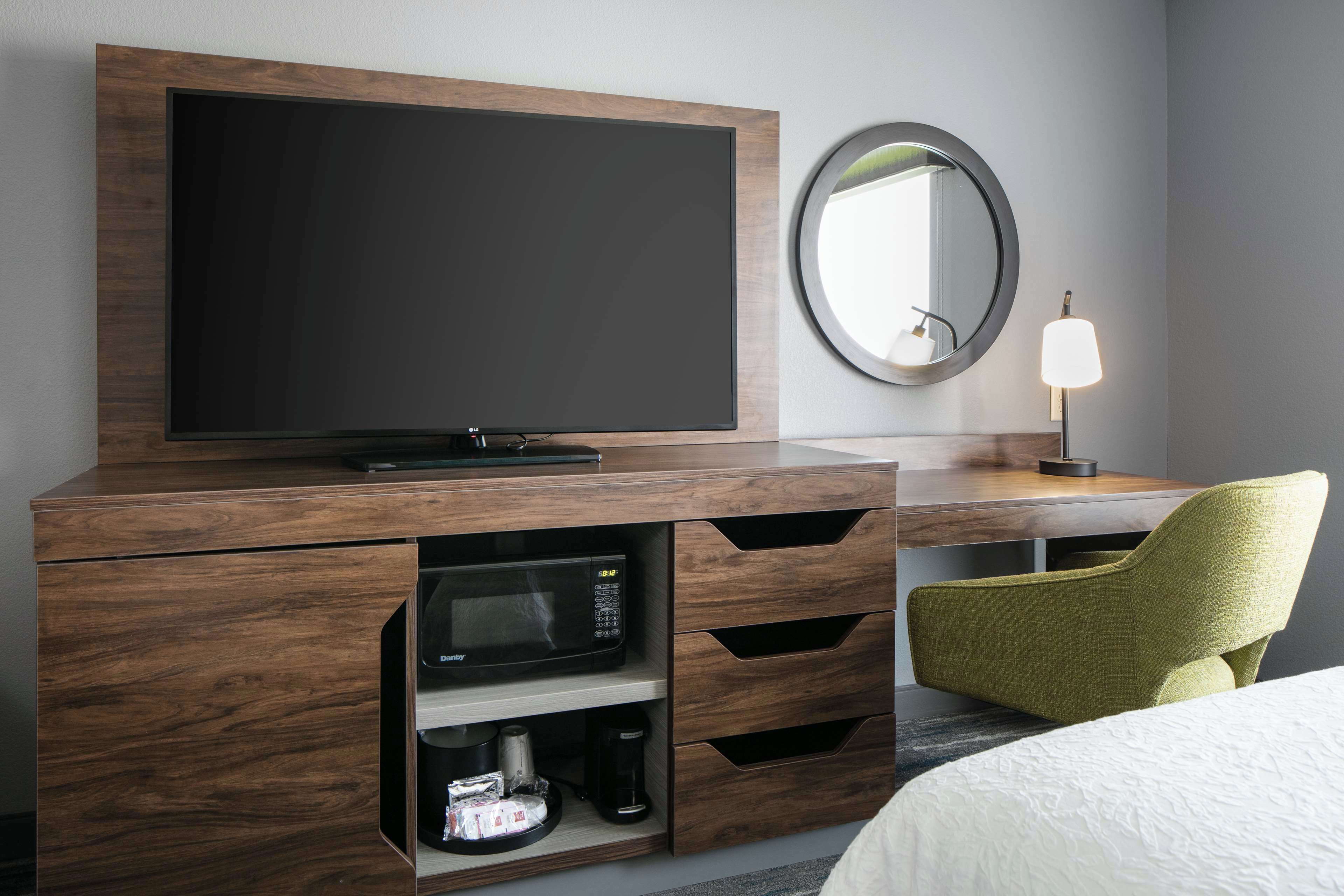 Ein Hotelzimmer mit einem Holz-Fernsehschrank, einem Flachbildfernseher, einer Mikrowelle, einer Kaffeemaschine, einem grünen Sessel, einem runden Spiegel und einer Nachttischlampe.
