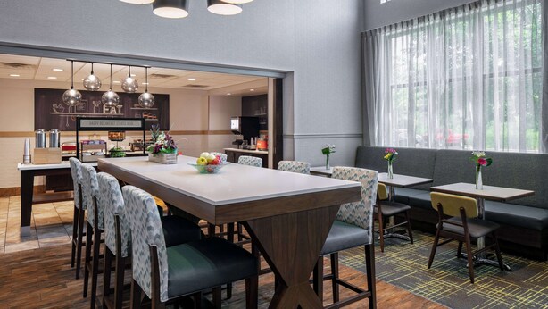 A modern dining area with a central table, surrounding chairs, and a sofa. There are pendant lights above the table and a large window with sheer curtains.