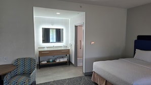 A hotel room with a bed, a desk, a chair, a bathroom with a sink and mirror, and a shower.