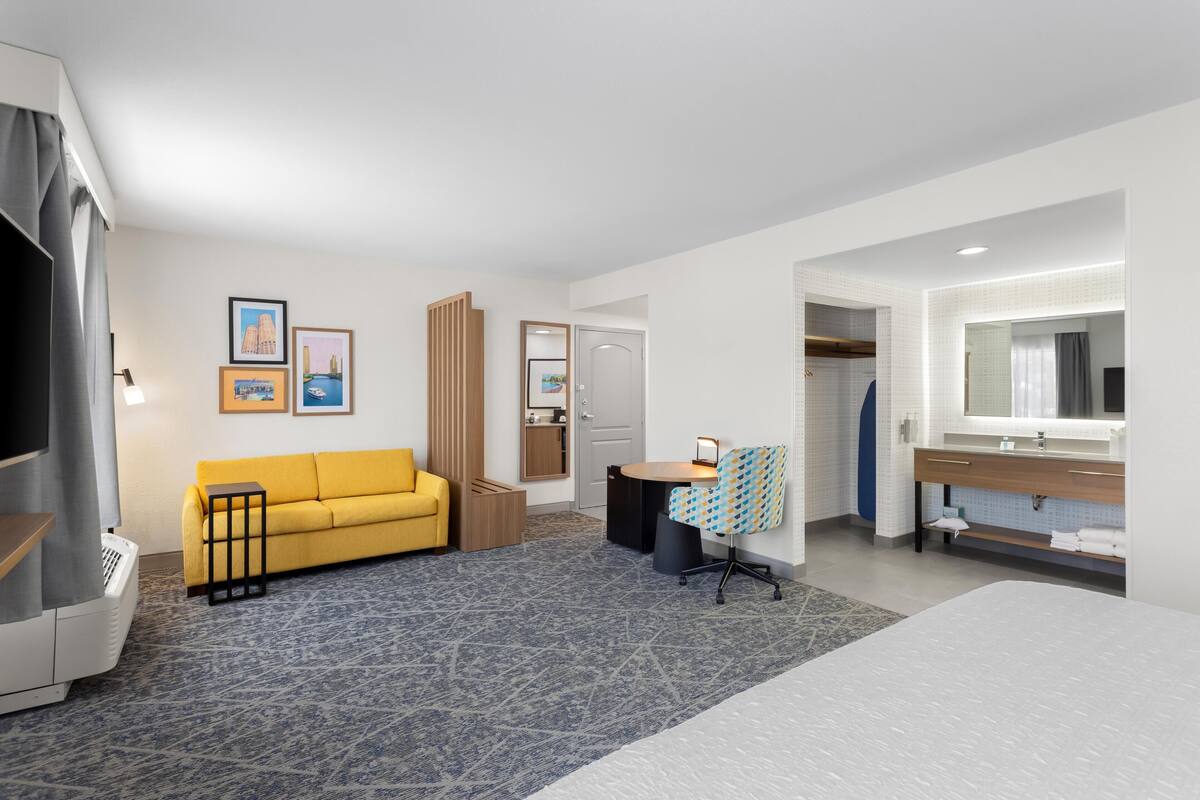 A hotel room with a bed, a sofa, a desk with a chair, a bathroom with a mirror, and framed pictures on the wall.