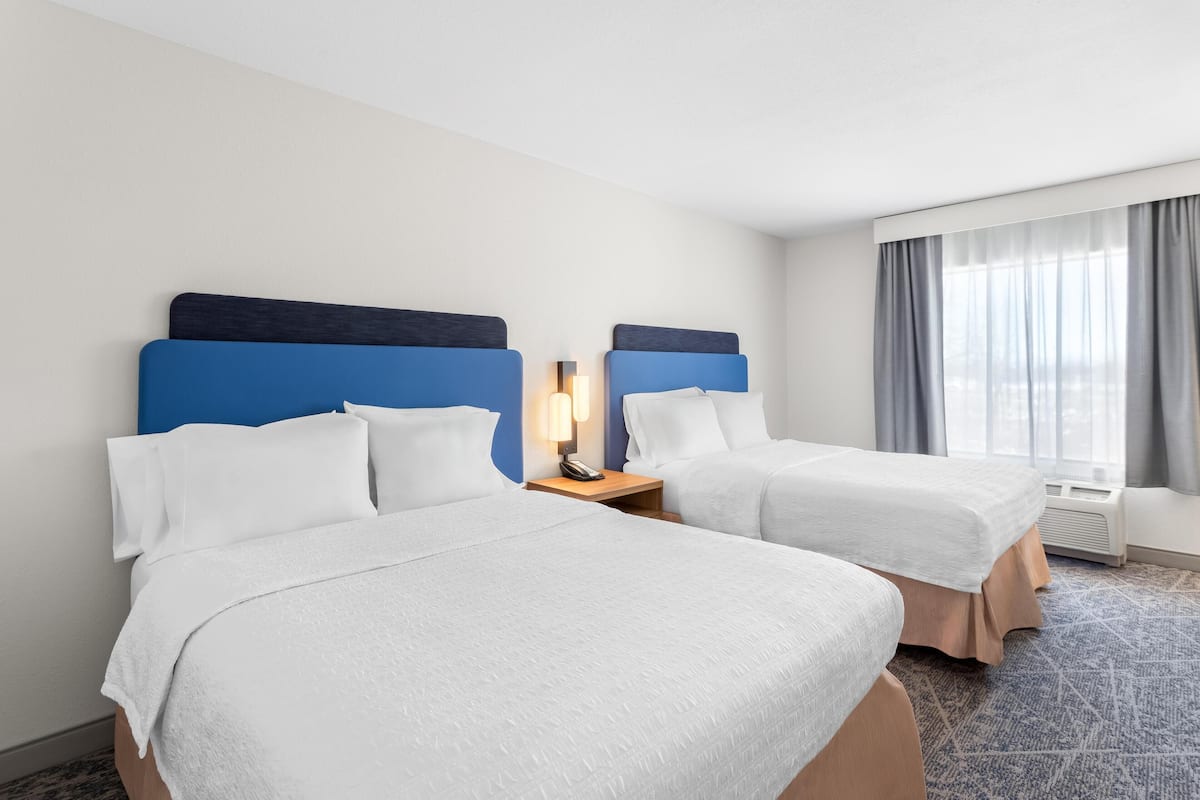 A hotel room with two beds, a nightstand with a lamp, a window with curtains, and a patterned carpet.
