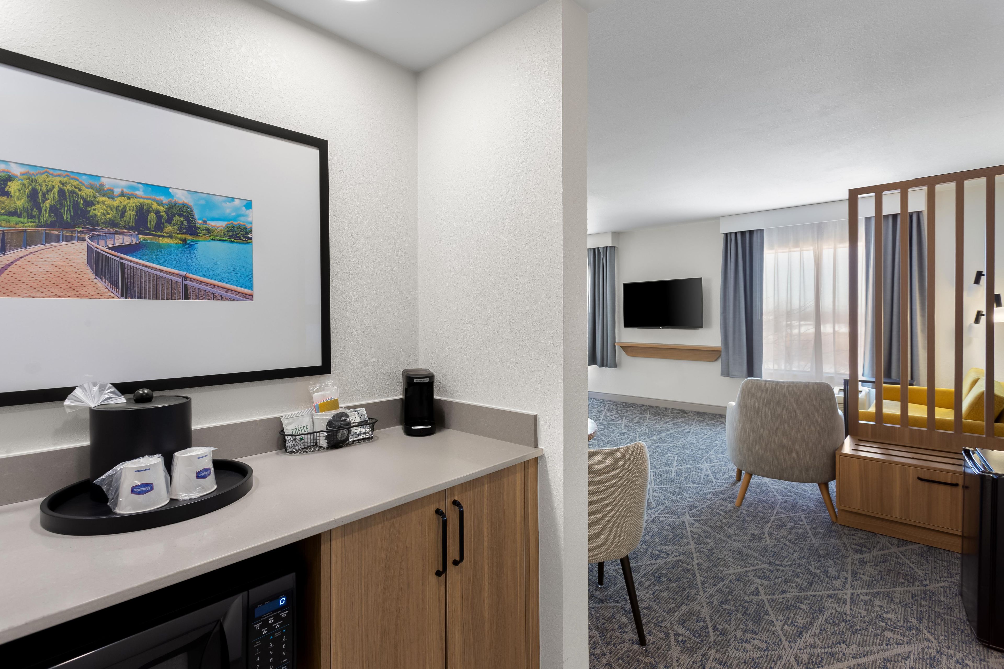 Habitación de hotel con un ventanal grande, televisión de pantalla plana, cafetera, microondas, una pequeña mesa y zona de estar con una silla y un sofá.