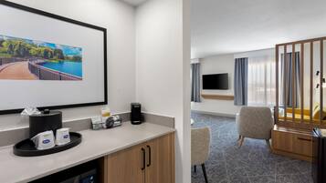 Habitación de hotel con un ventanal grande, televisión de pantalla plana, cafetera, microondas, una pequeña mesa y zona de estar con una silla y un sofá.